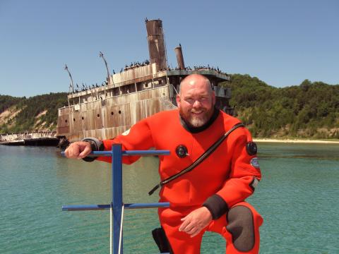 man in an orange dive suit in the water posing before an old ship