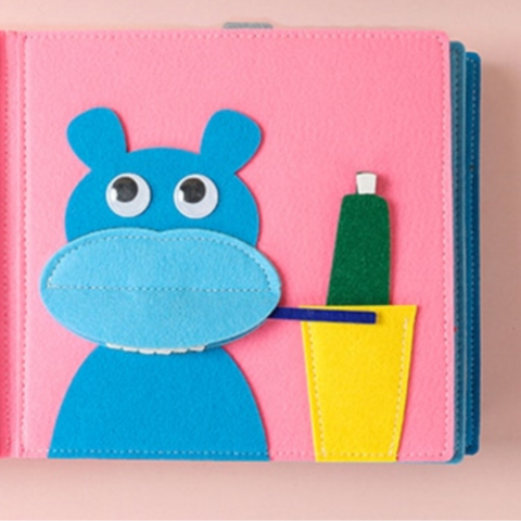 Pink background, blue hippopotamus focusing on brushing teeth (made out of felt pieces)